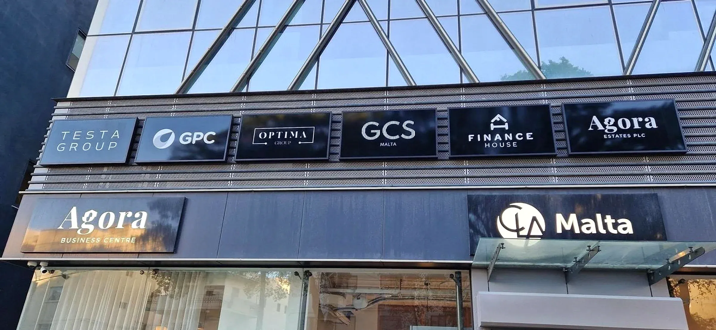Row of six dark tenant name panels above two larger illuminated signs on a glazed building frontage