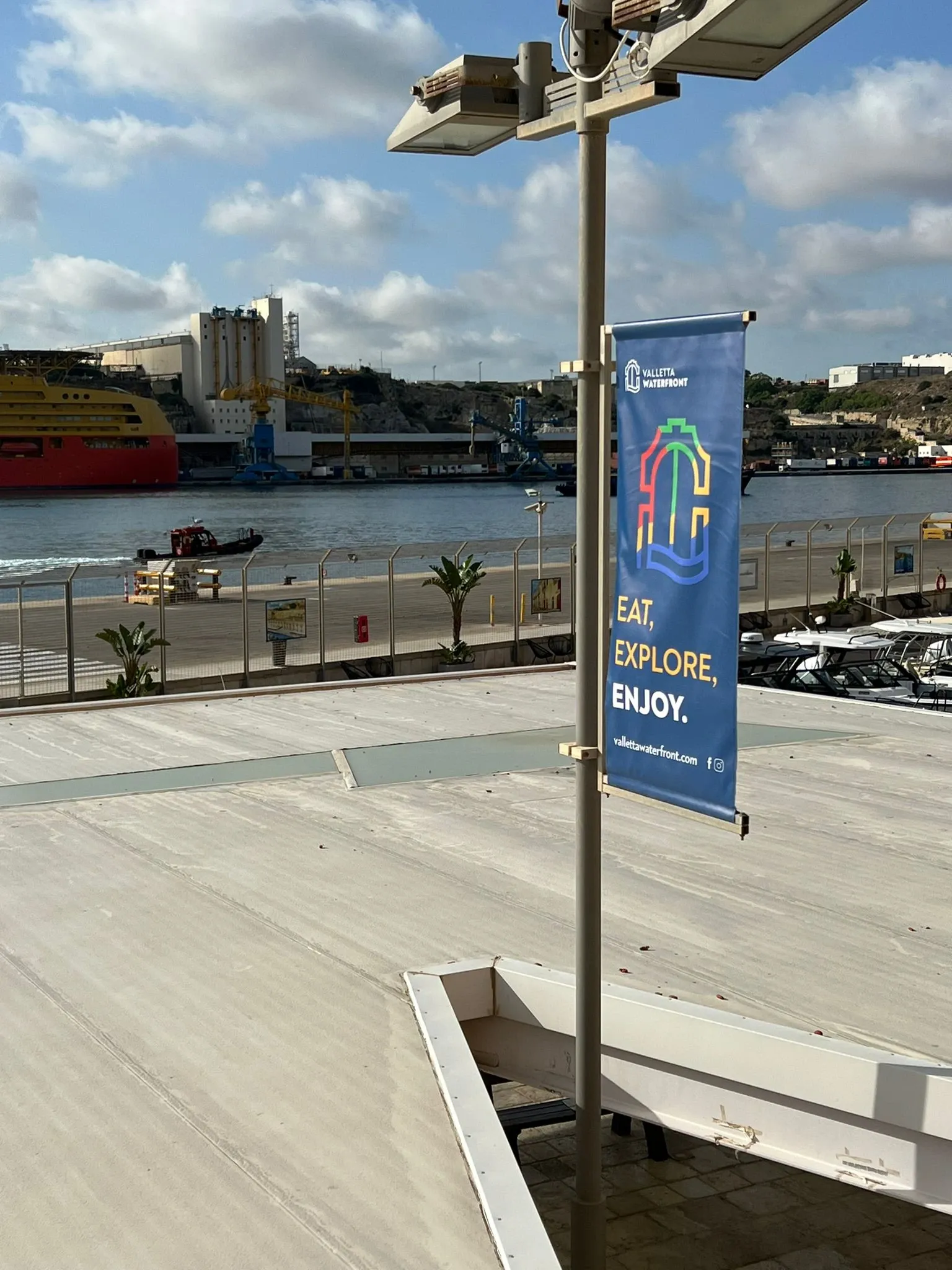 Blue lamp-post banner on a waterfront promenade with a harbour, moored ship and quay behind