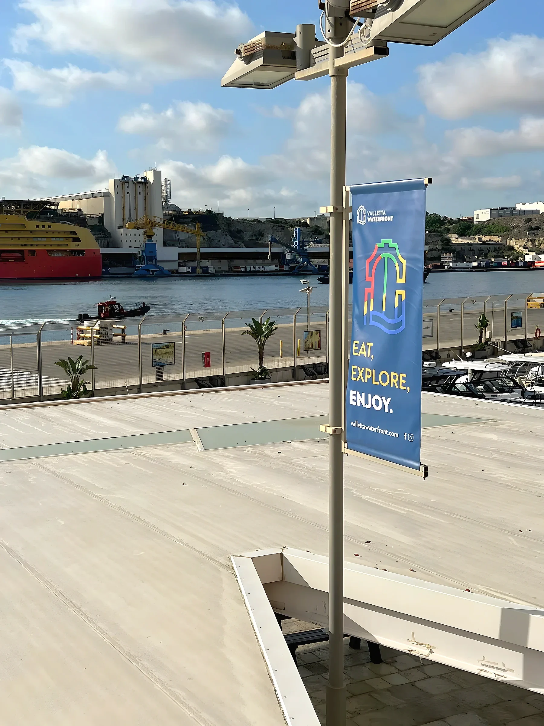 Blue lamp-post banner on a waterfront promenade with a harbour, moored ship and quay behind