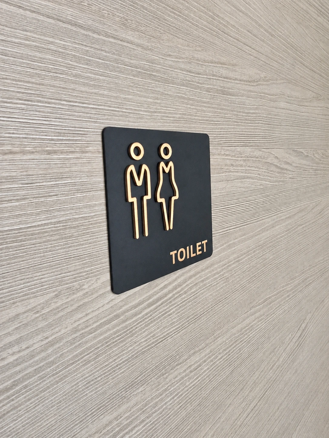 Black square toilet sign with raised gold figures and lettering on a pale timber wall