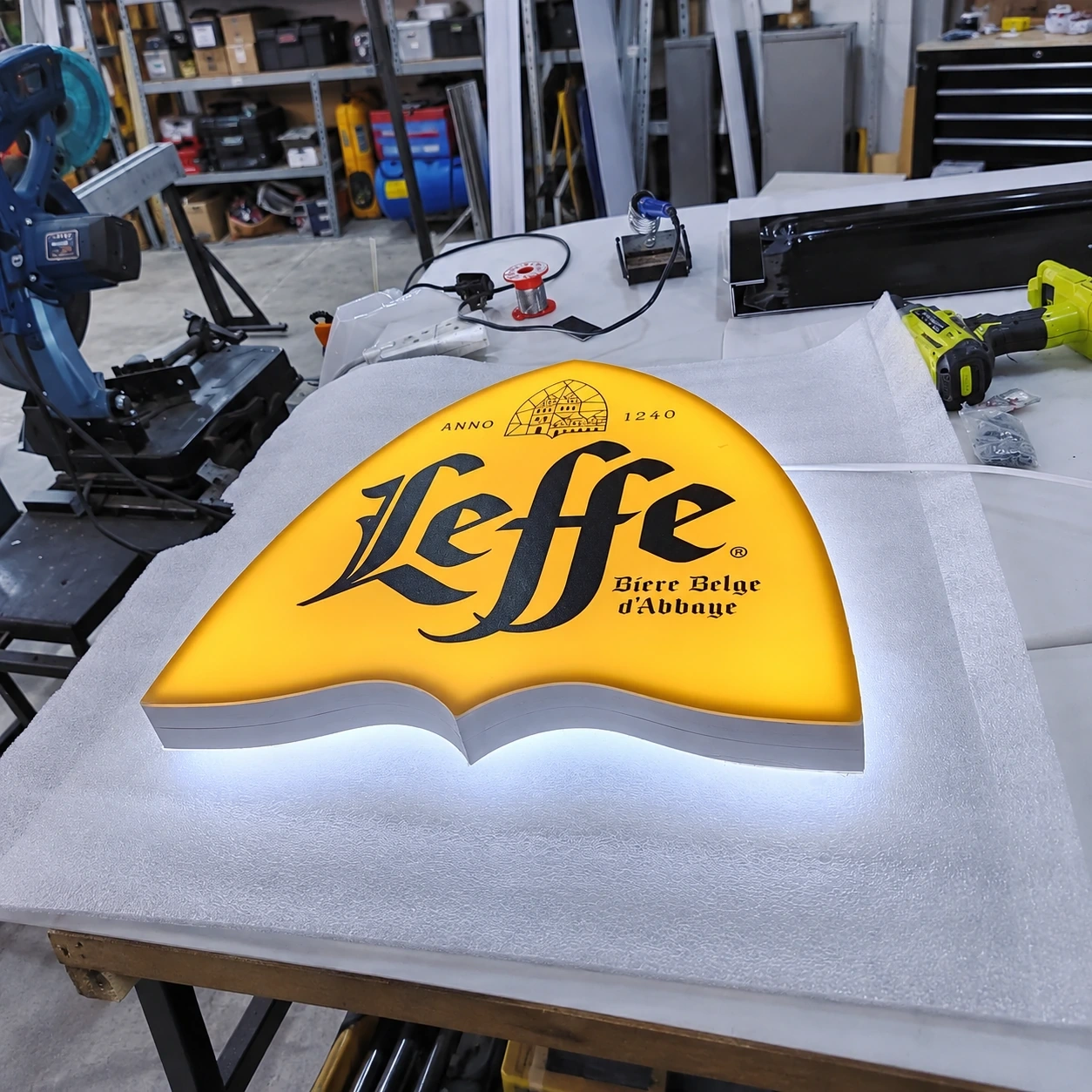 Shaped halo-lit sign lying face up on a workshop bench, its yellow face glowing against the light behind it