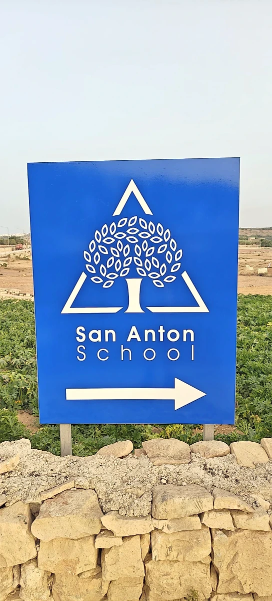 Blue directional sign with a white tree emblem and an arrow, set on a rubble wall beside fields