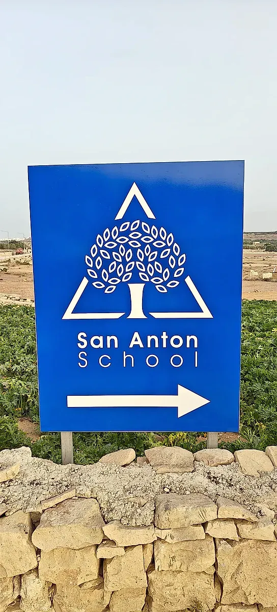 Blue directional sign with a white tree emblem and an arrow, set on a rubble wall beside fields