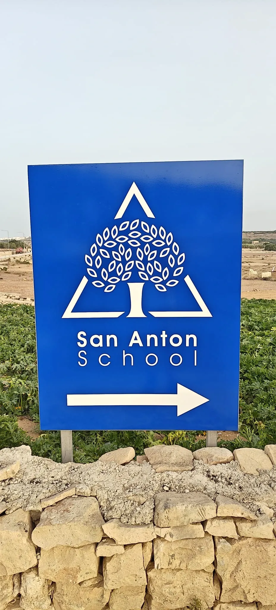 Blue directional sign with a white tree emblem and an arrow, set on a rubble wall beside fields
