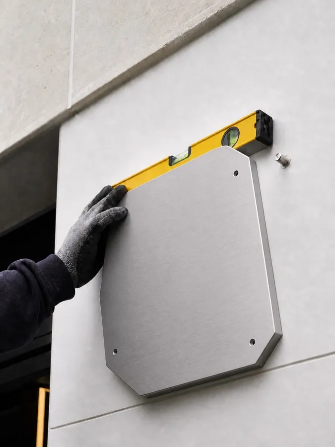 Gloved hand levelling a blank asymmetrical aluminium plate against a fascia