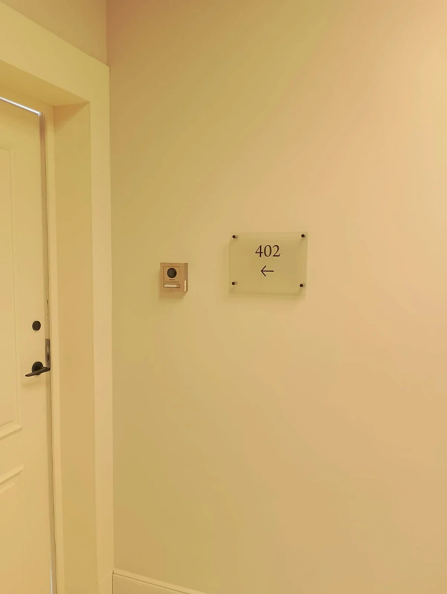 Small acrylic room number plate with an arrow, fixed to a corridor wall on standoffs