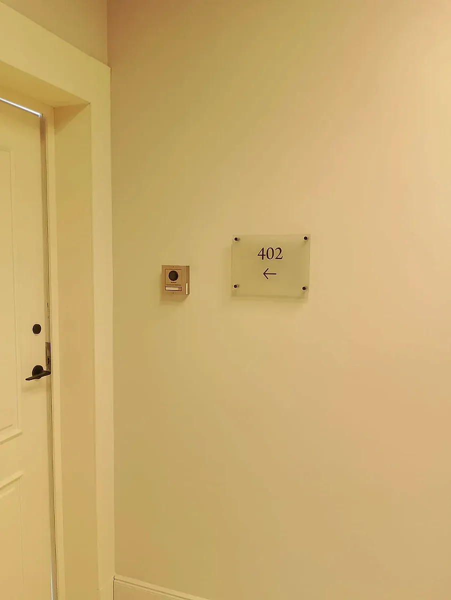 Small acrylic room number plate with an arrow, fixed to a corridor wall on standoffs
