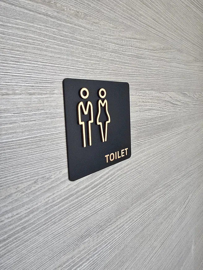 Black square toilet sign with raised gold figures and lettering on a pale timber wall