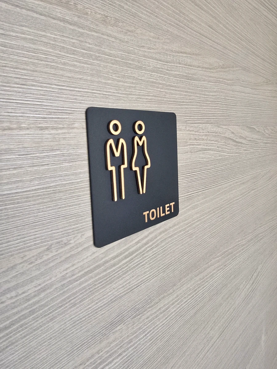 Black square toilet sign with raised gold figures and lettering on a pale timber wall