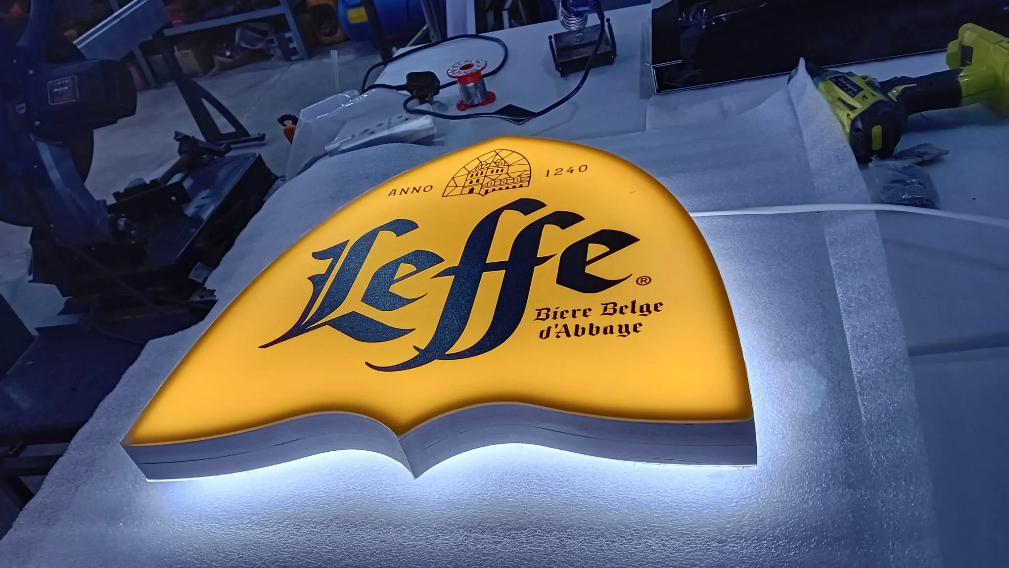 Shaped halo-lit sign lying face up on a workshop bench, its yellow face glowing against the light behind it