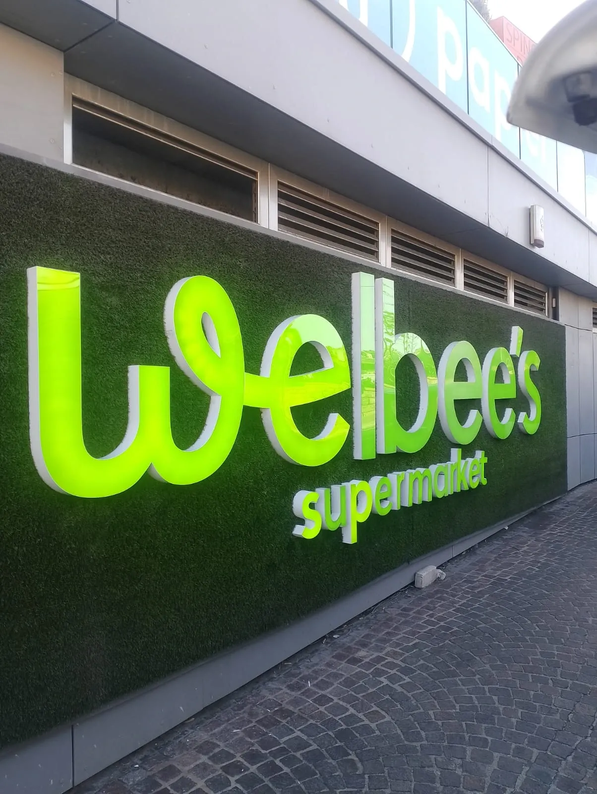 Illuminated green built-up letters spelling Welbee's supermarket, mounted across an artificial grass shopfront wall