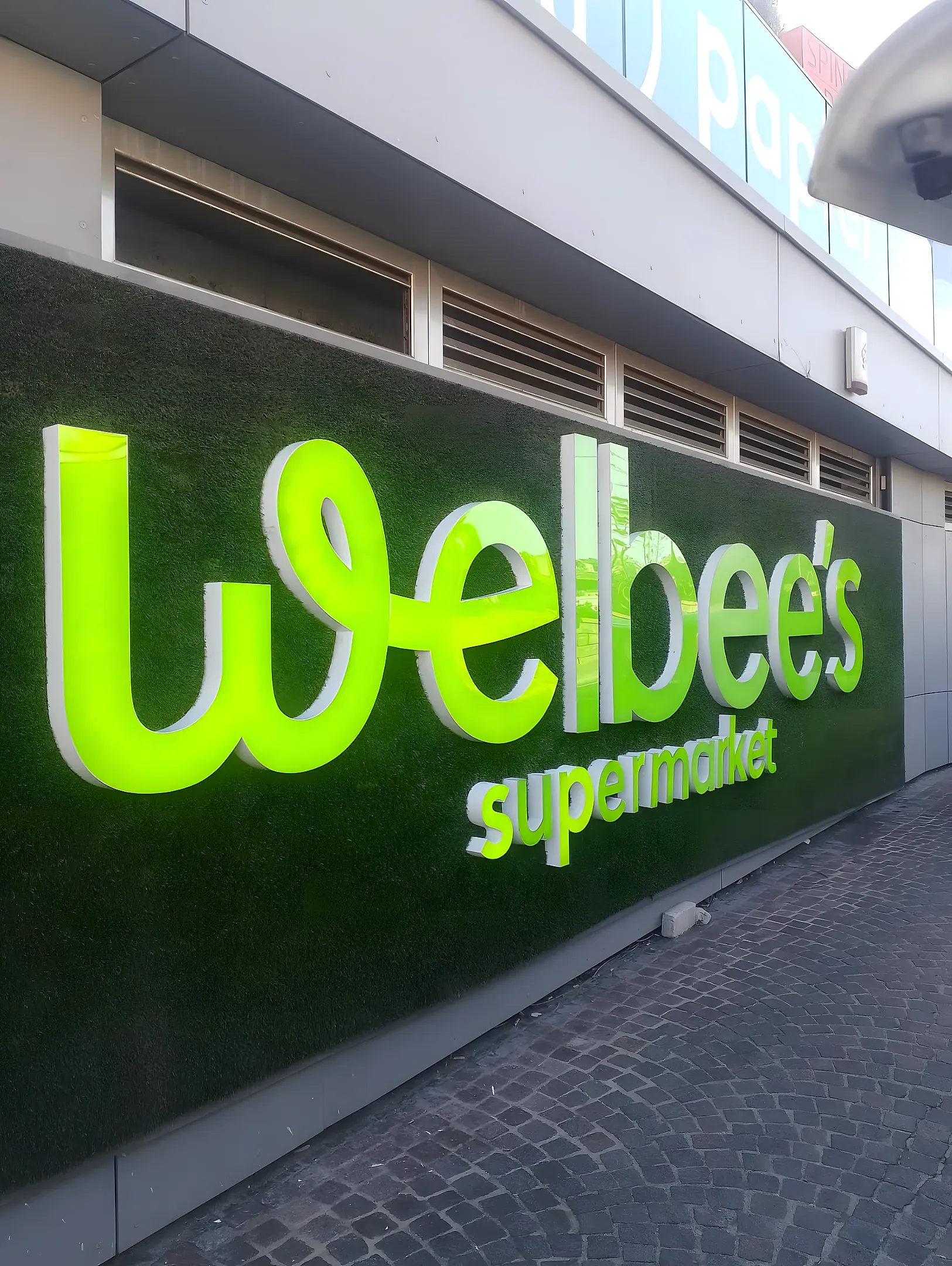 Illuminated green built-up letters spelling Welbee's supermarket, mounted across an artificial grass shopfront wall