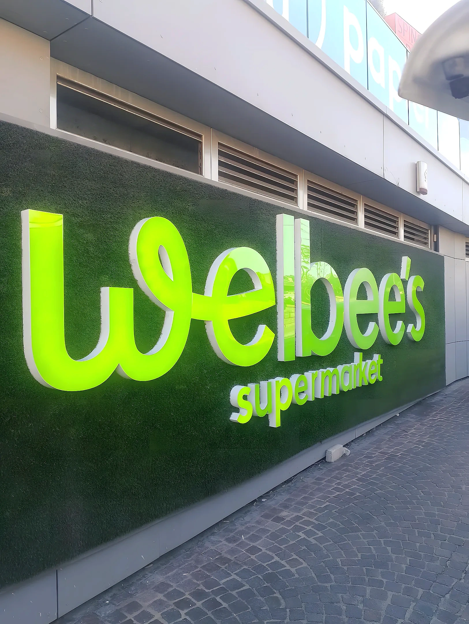 Illuminated green built-up letters spelling Welbee's supermarket, mounted across an artificial grass shopfront wall