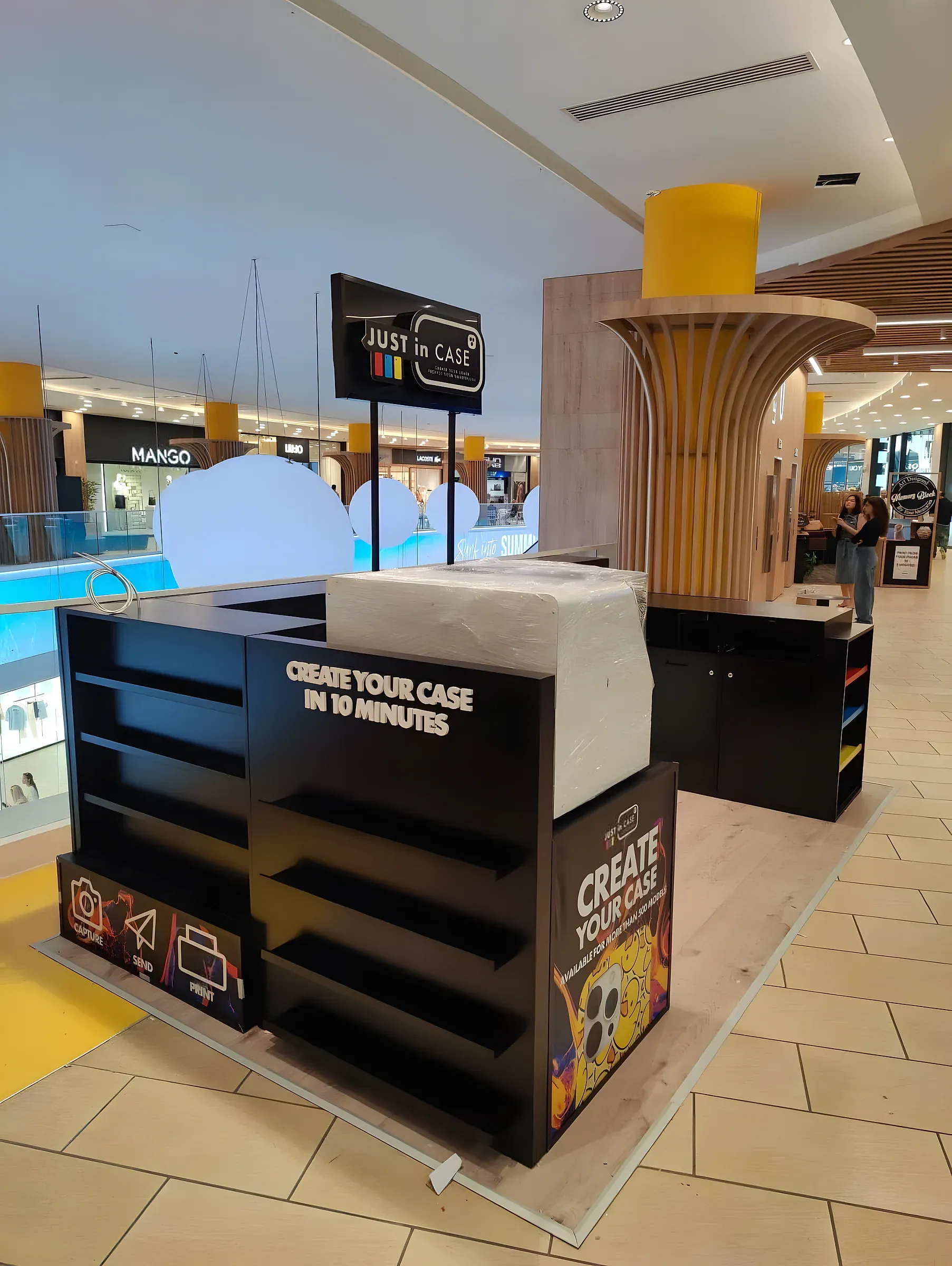 Black branded retail kiosk with open shelving and printed panels, standing on a shopping centre floor