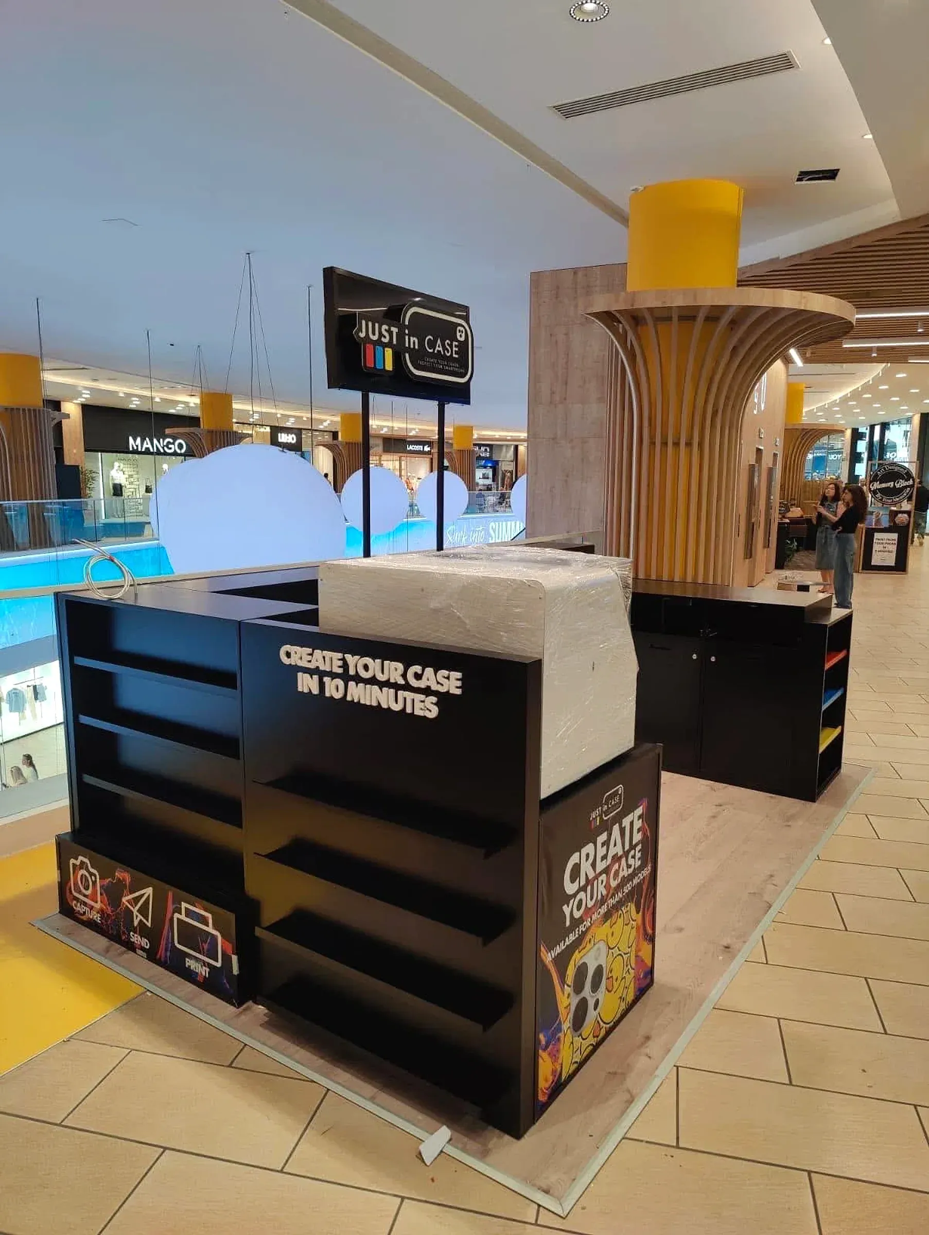 Black branded retail kiosk with open shelving and printed panels, standing on a shopping centre floor