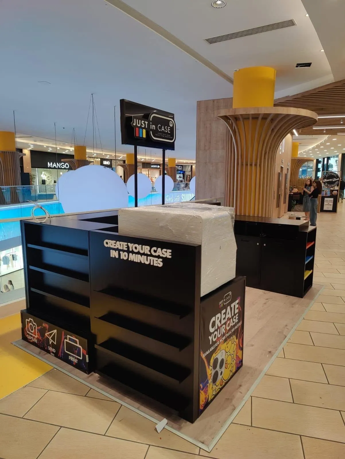 Black branded retail kiosk with open shelving and printed panels, standing on a shopping centre floor