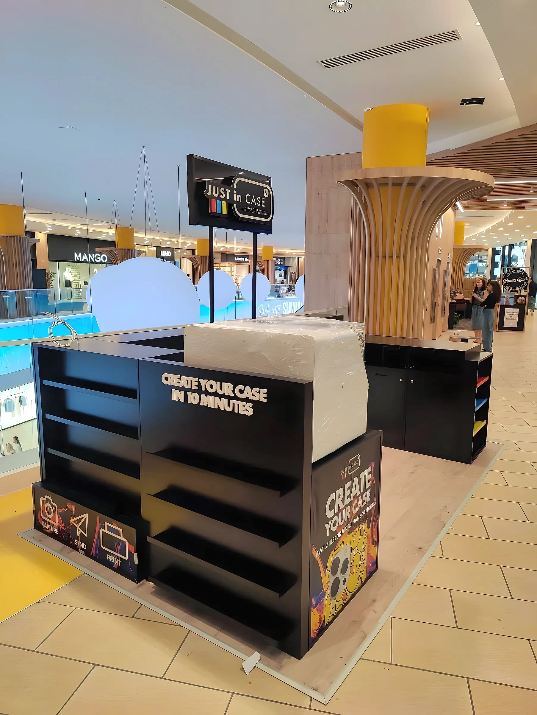 Black branded retail kiosk with open shelving and printed panels, standing on a shopping centre floor