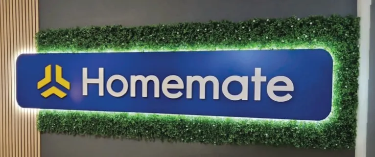 Backlit shopfront sign with raised three-dimensional lettering on a deep blue panel, framed by a dense green foliage border