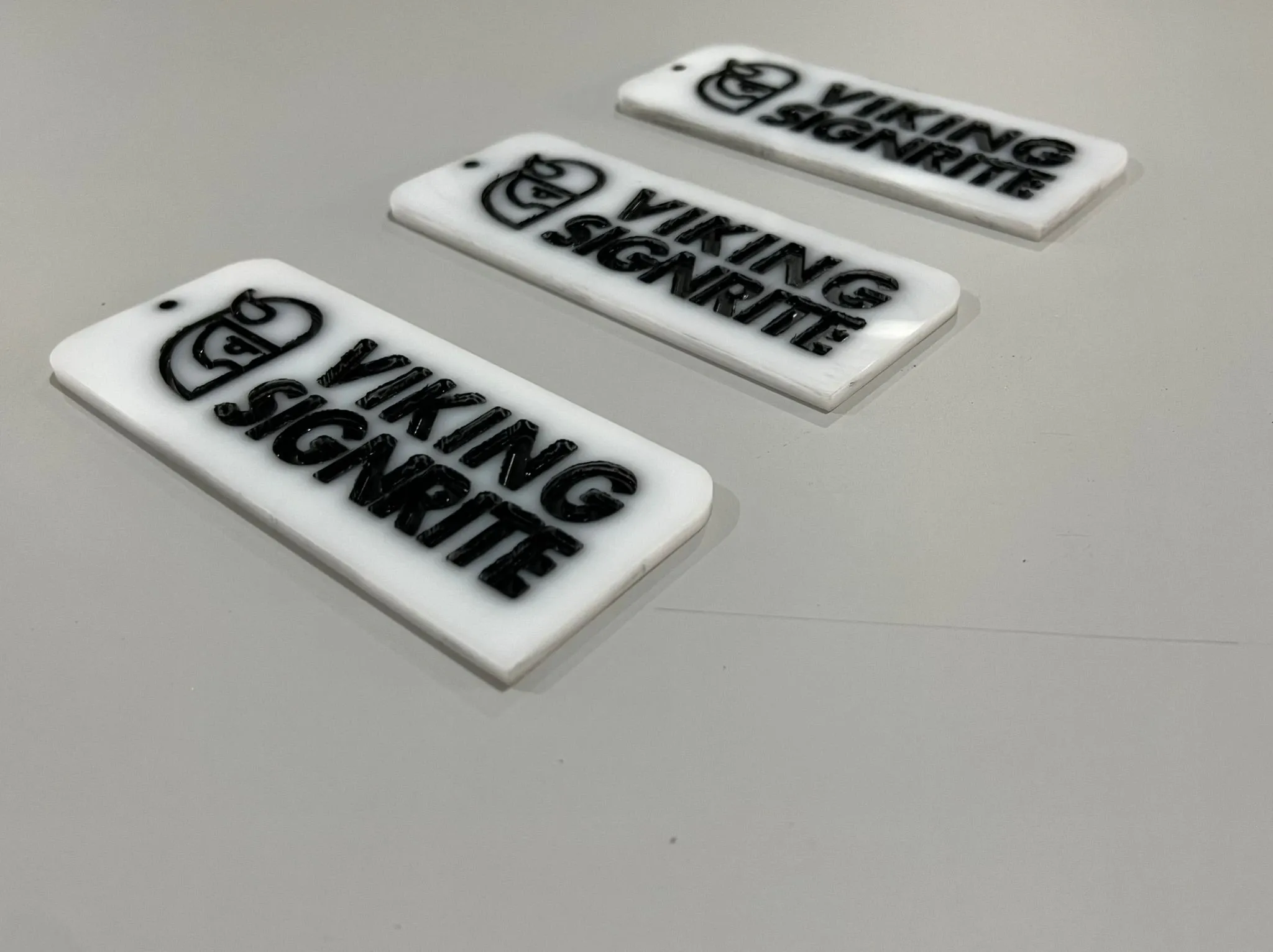 Three engraved white acrylic tags carrying the Viking Signrite logo, laid out in a diagonal row on a pale surface