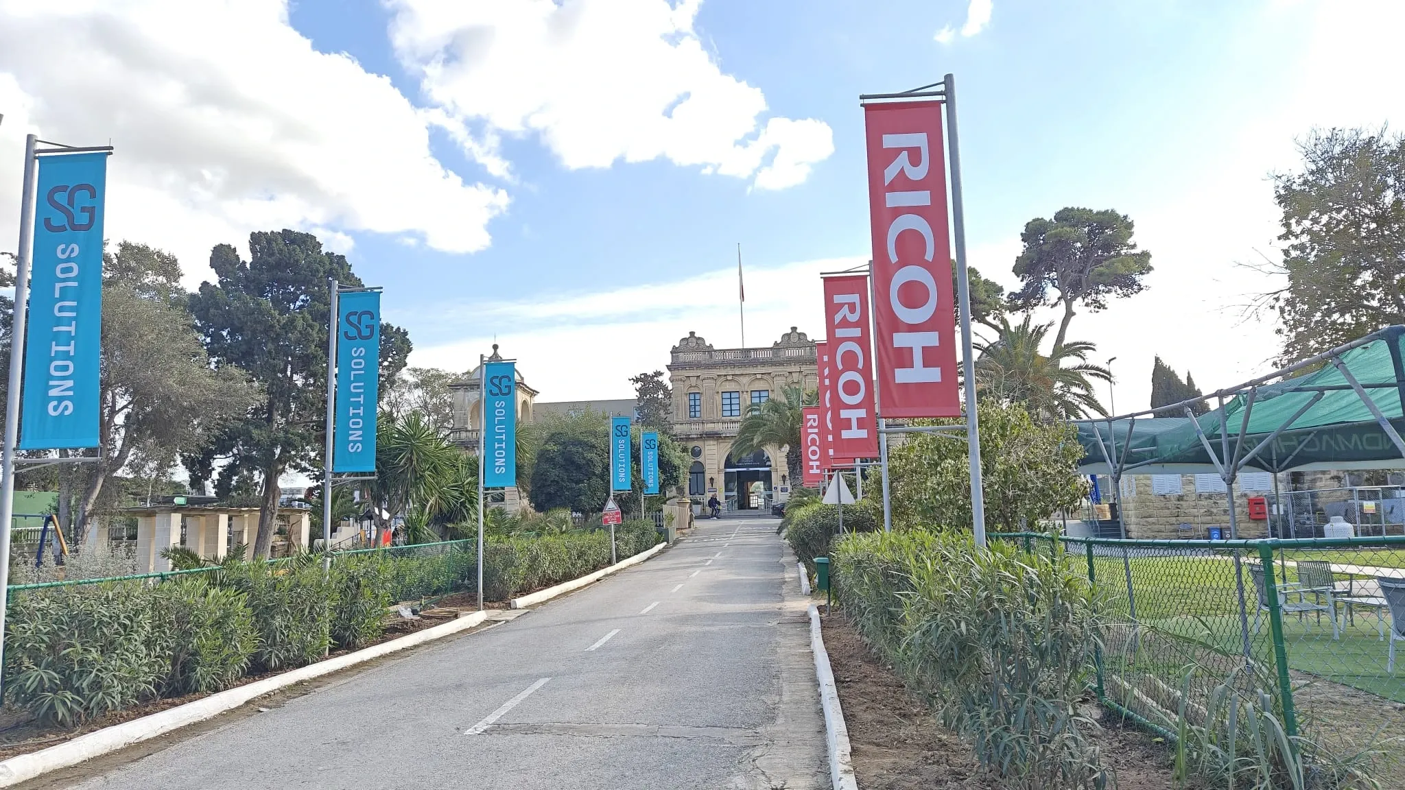 Run of blue and red printed banner flags on poles lining a driveway up to a historic stone building