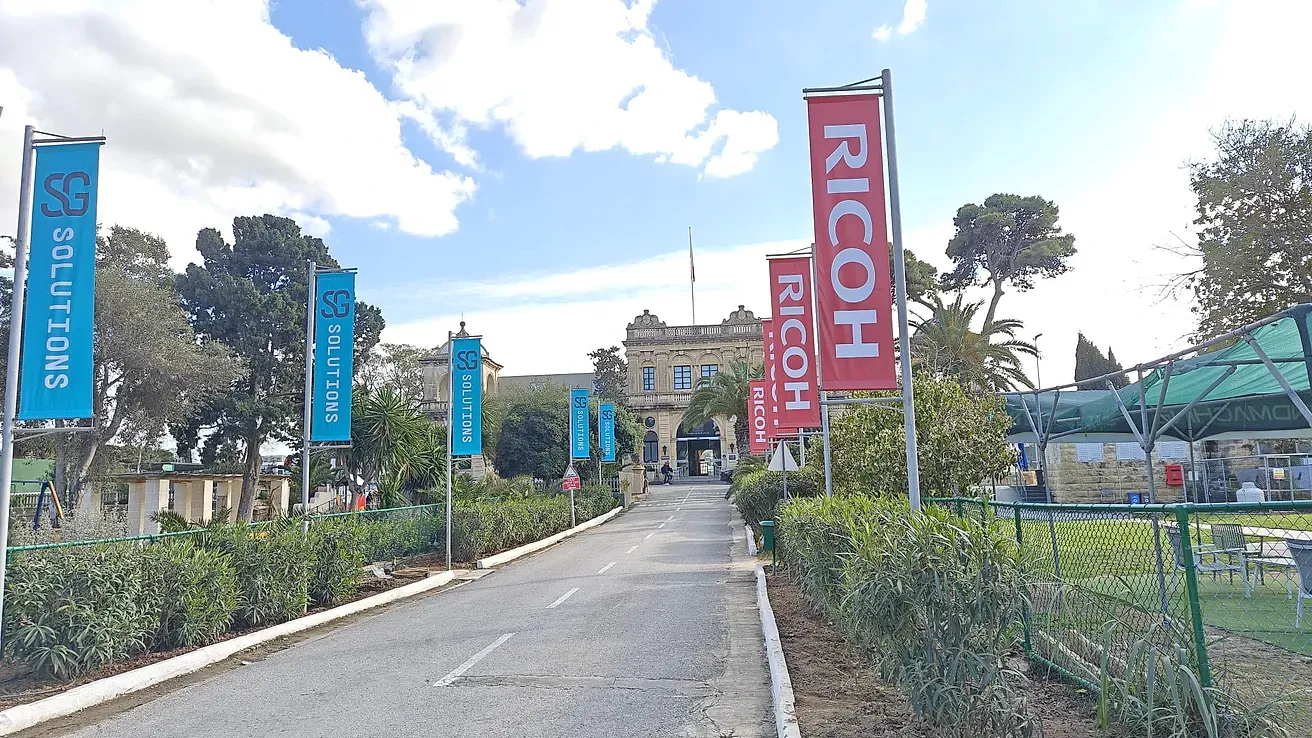 Run of blue and red printed banner flags on poles lining a driveway up to a historic stone building