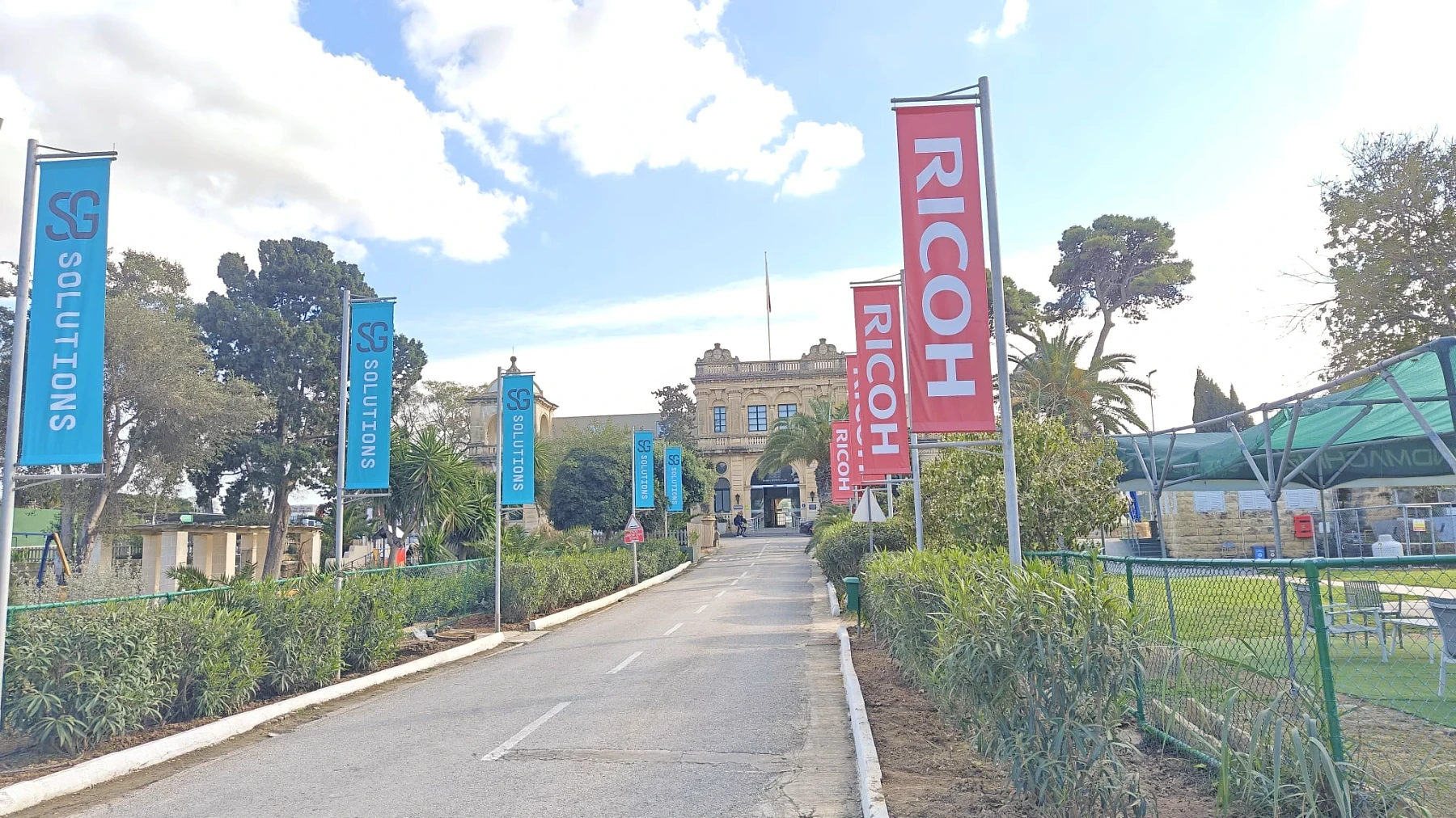 Run of blue and red printed banner flags on poles lining a driveway up to a historic stone building
