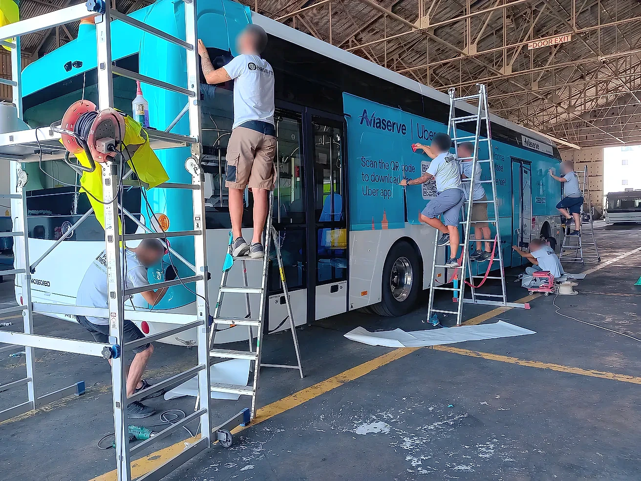 Fitters on ladders applying printed vinyl along the side of a bus inside a depot