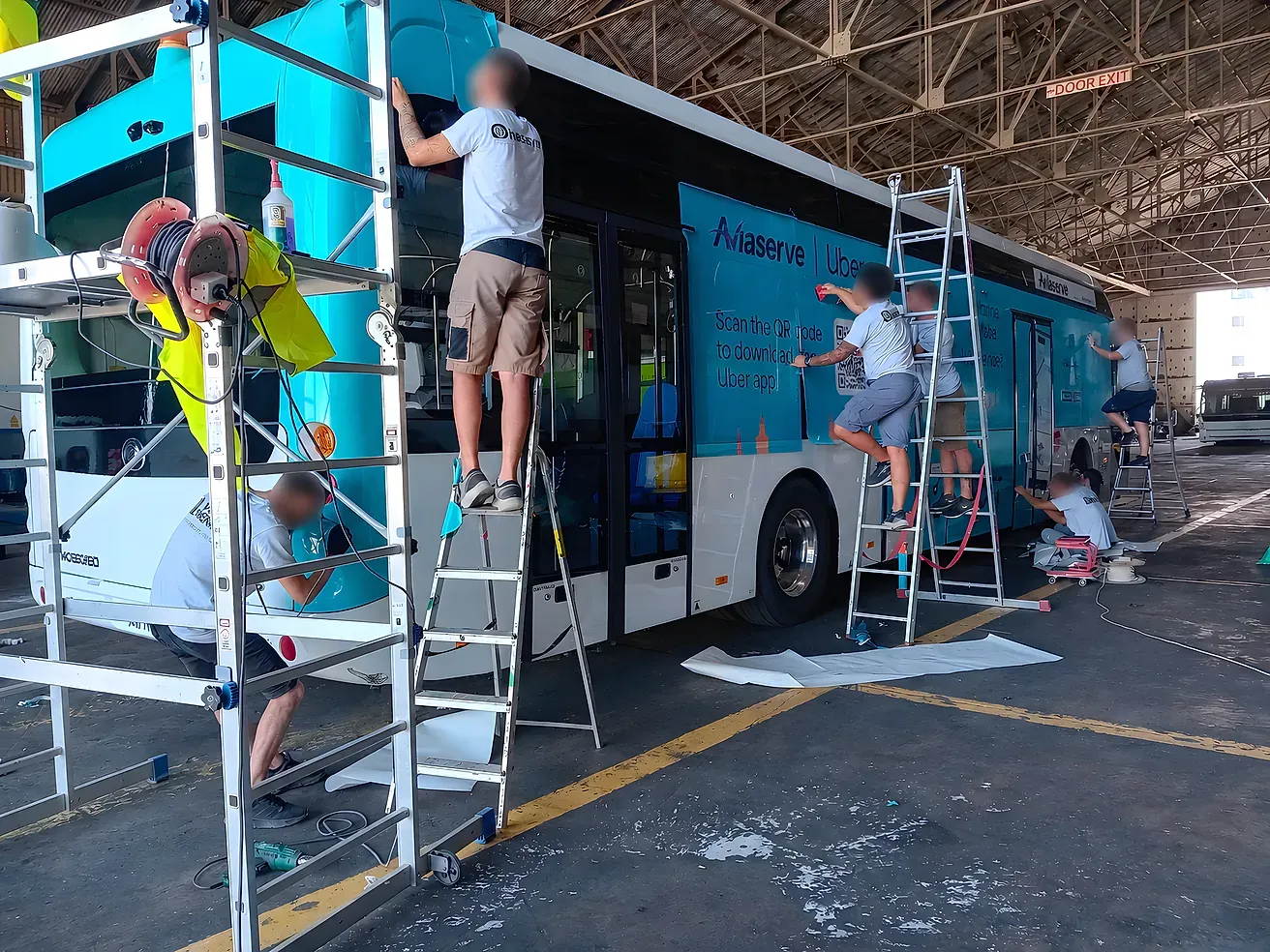 Fitters on ladders applying printed vinyl along the side of a bus inside a depot