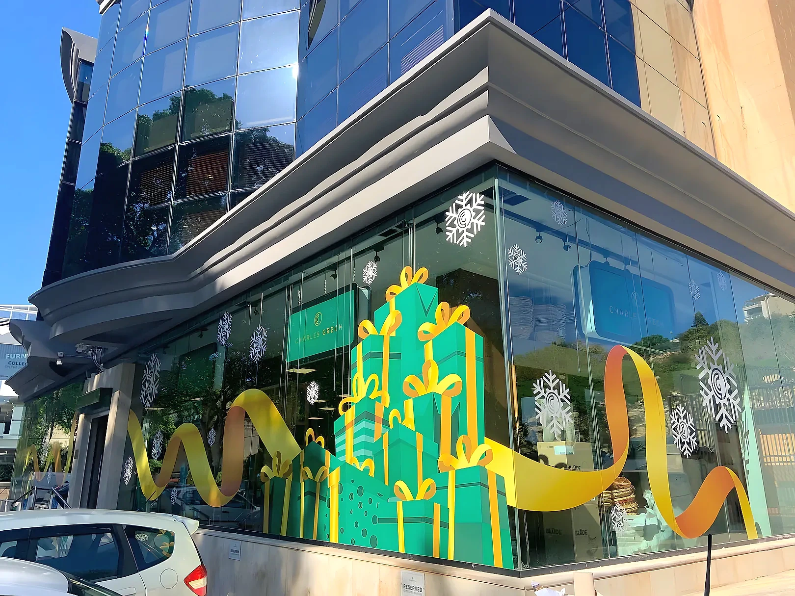 Glazed corner shopfront with printed window graphics of stacked green gift boxes, gold ribbon and white snowflakes