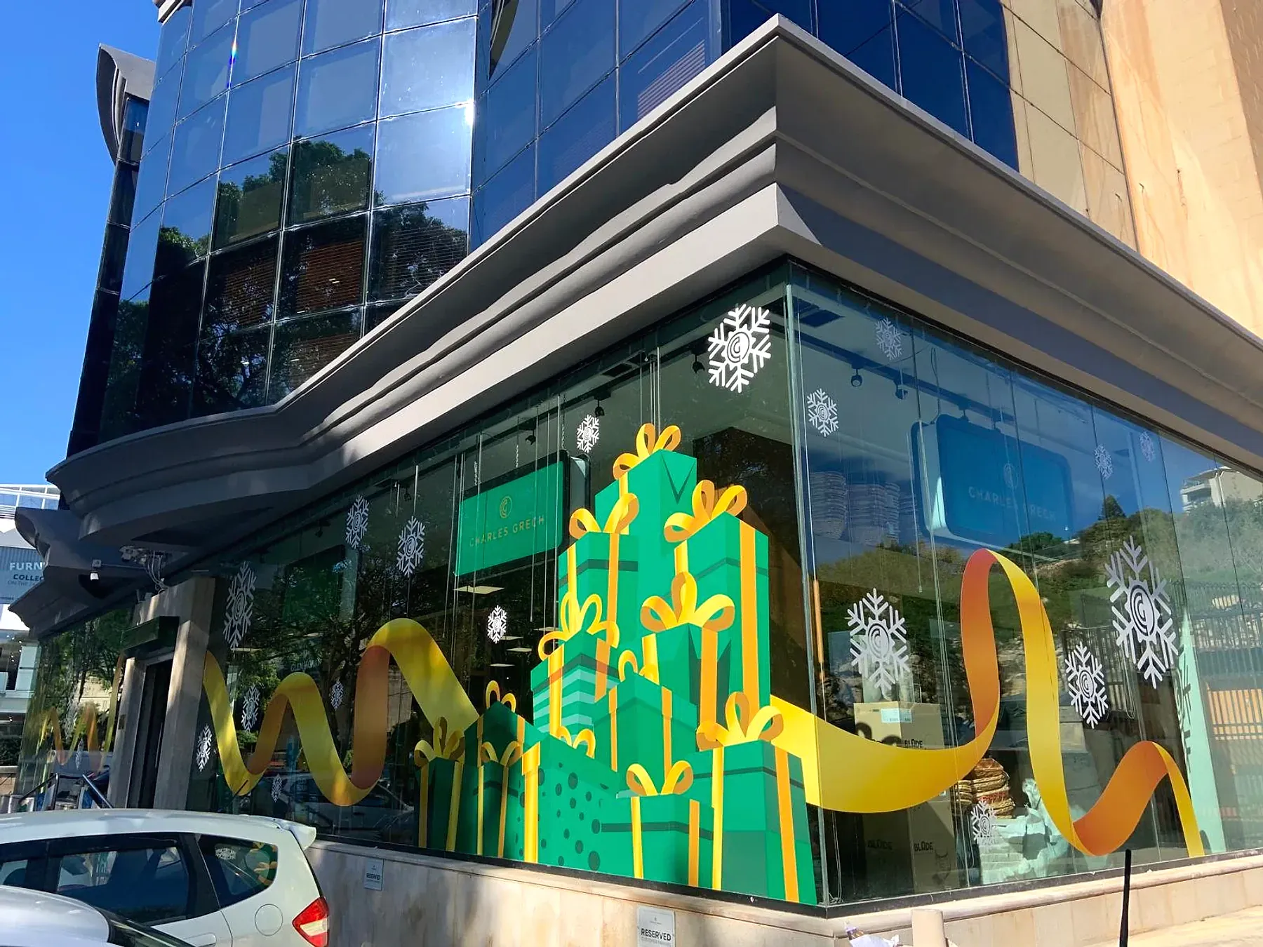 Glazed corner shopfront with printed window graphics of stacked green gift boxes, gold ribbon and white snowflakes