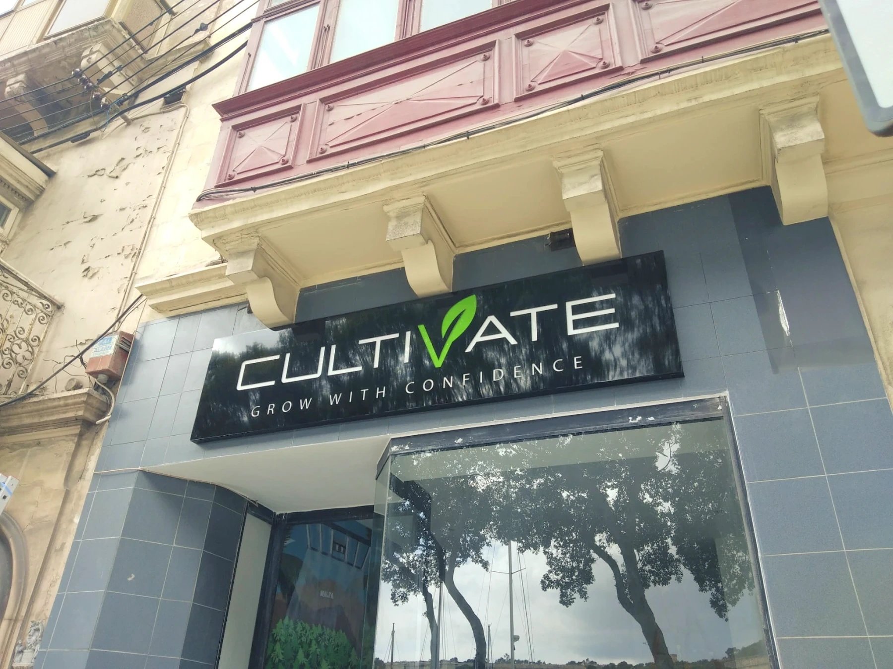 Black fascia sign with built-up white lettering and a green leaf detail, mounted above a tiled shopfront below a traditional balcony