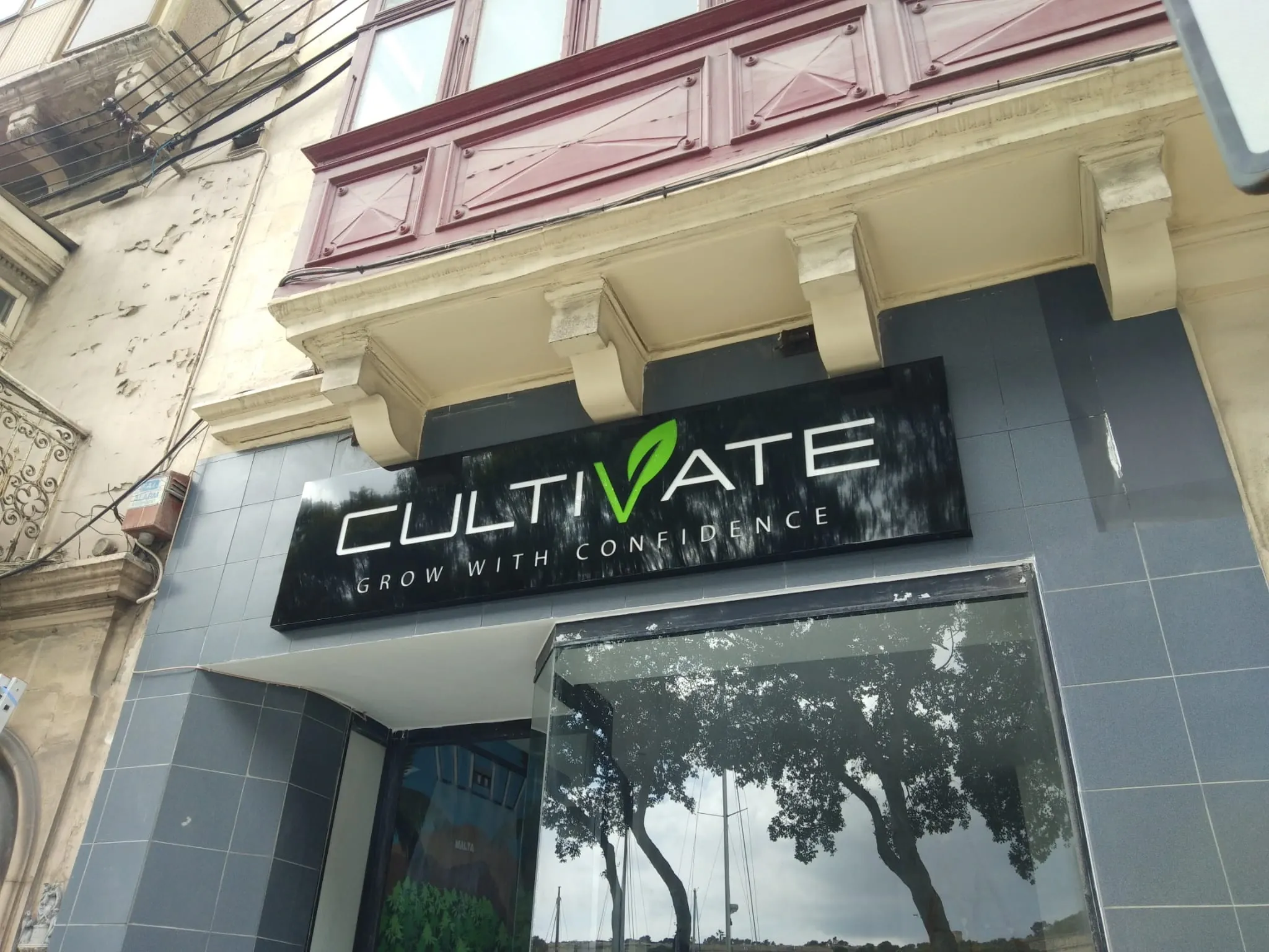 Black fascia sign with built-up white lettering and a green leaf detail, mounted above a tiled shopfront below a traditional balcony