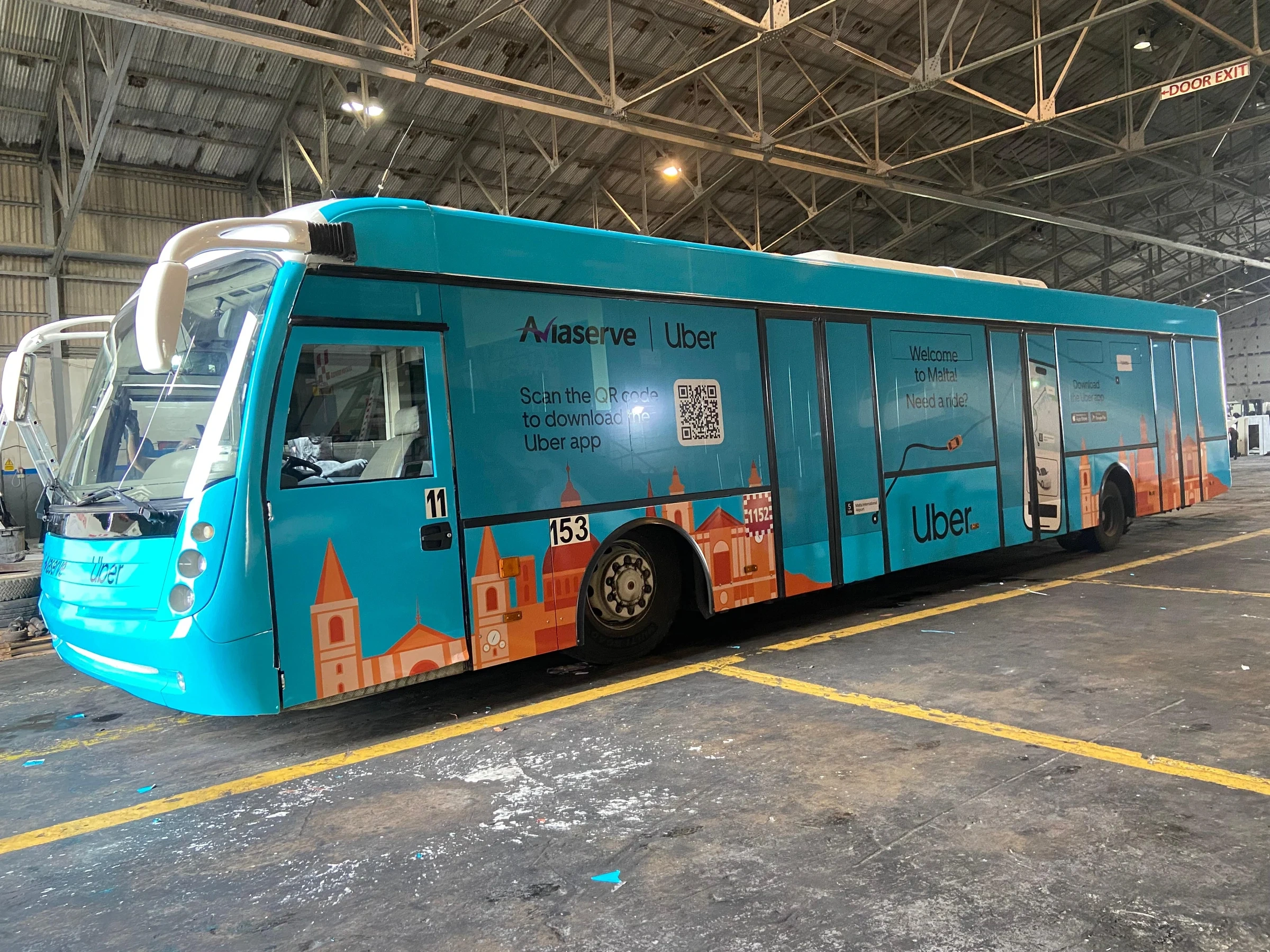 Turquoise airport bus with a full printed wrap showing a Maltese skyline, parked inside a depot