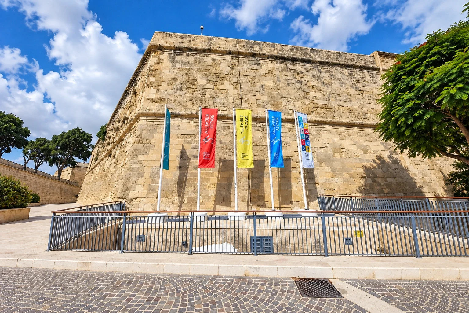 Five coloured banner flags on angled poles against a limestone bastion wall under a blue sky
