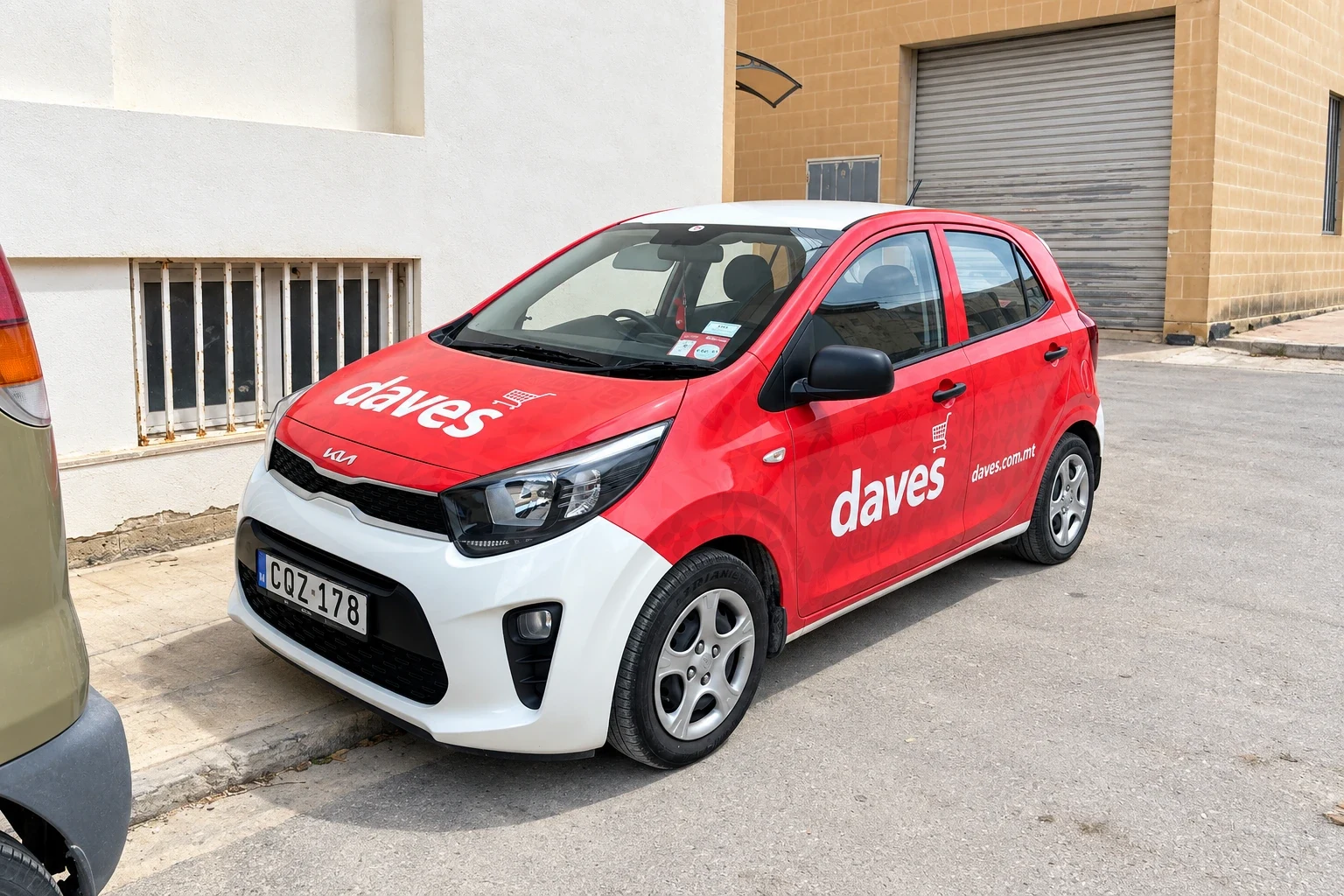 Red and white wrapped city car with a brand name on the bonnet and door, parked by a warehouse