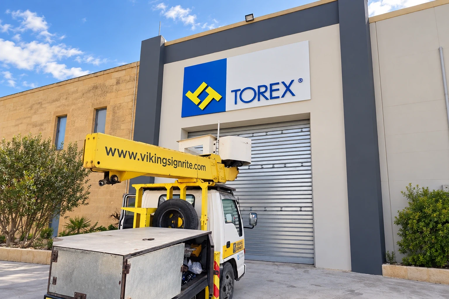 Blue and yellow logo panel with built-up lettering high on an industrial facade, with a cherry picker below