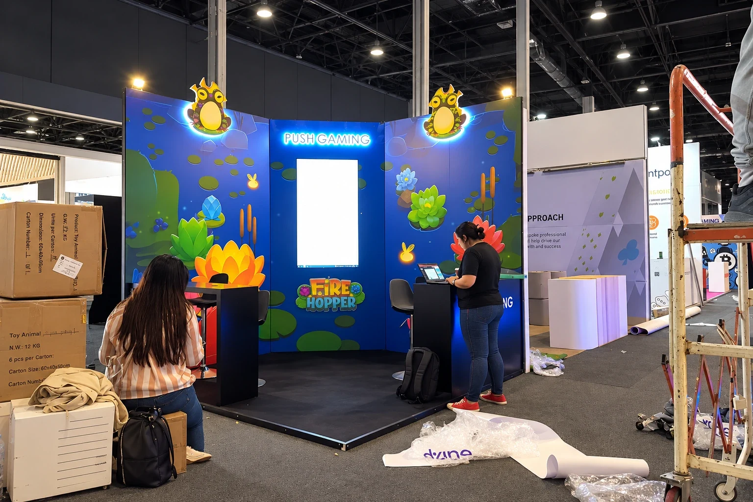 Illustrated gaming exhibition stand with lit character panels and a central lightbox being set up on a show floor