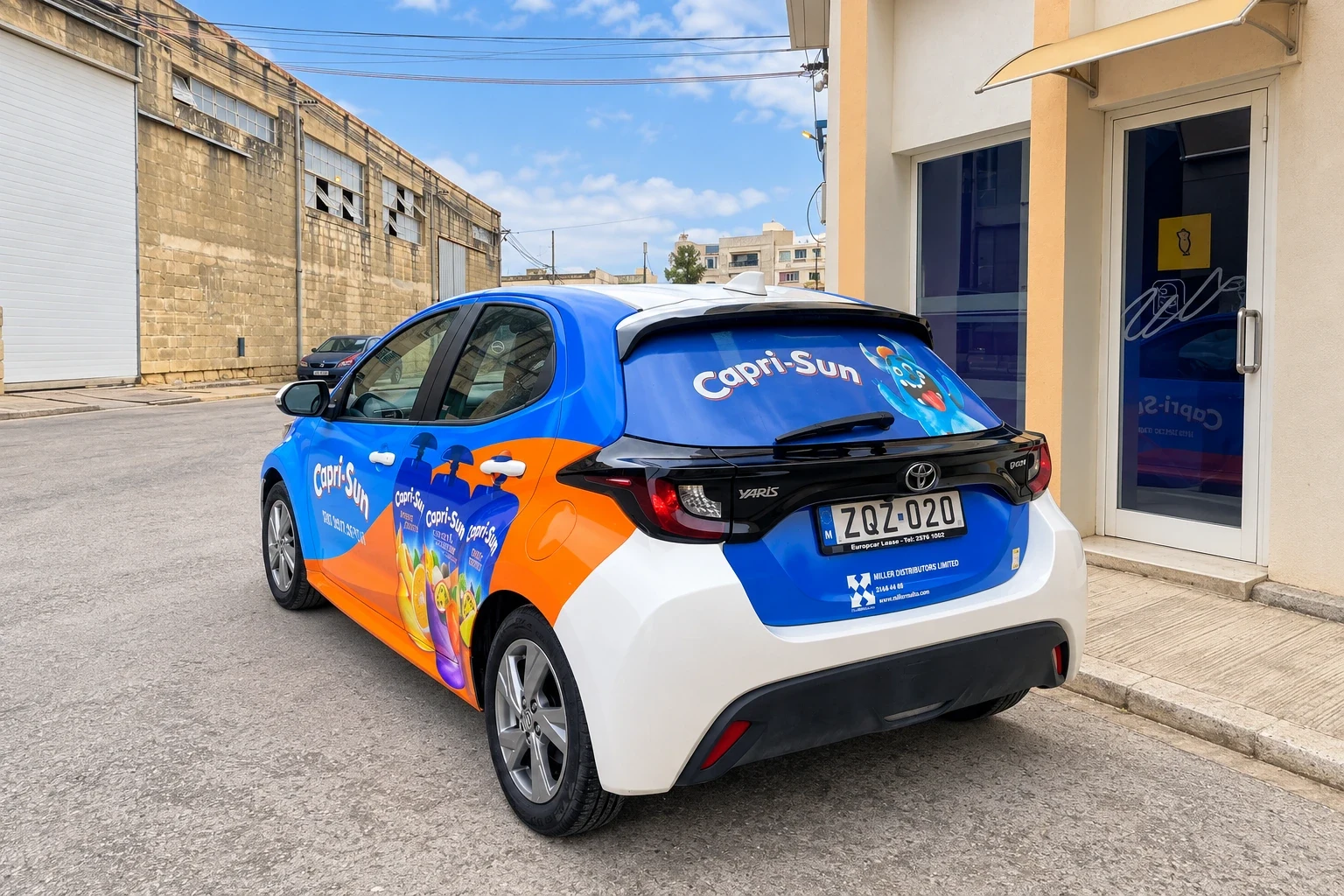 Blue and orange wrapped hatchback with a printed rear screen, parked outside a workshop